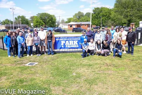 A group of people gathered around a sign reading "Rotary Club of Paducah Park-A-Palooza" on a grassy area.